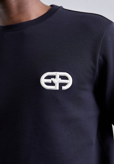 Tengerészkék pamut pulóver kerek nyakkal, fehér hímzett logóval a mellrészen. Sima textúrájú és testhezálló fazonú.