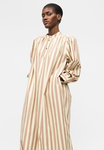Γυναίκα που φοράει μια άνετη μακριά φόρεμα με κάθετες ρίγες σε μπεζ και ανοιχτό καφέ, με μακριά μανίκια, ψηλό γιακά και κουμπωτό λαιμόκοψη, τα χέρια της στις τσέπες.