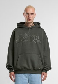 Hanorac cu glugă de culoare verde închis, cu un buzunar frontal. Inscripția "Nowhere is Now Here" în litere de culoare argintie, având manșete și tiv din tricot.