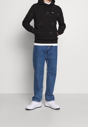 Sudadera negra con pequeños patrones de logo, combinada con jeans de mezclilla azul claro y zapatillas blancas. La sudadera cuenta con un bolsillo tipo canguro y cordones.