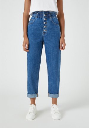 Femme portant un jean bleu taille haute avec boutons sur le devant, ourlets retroussés, associée à des baskets blanches et un haut blanc, debout devant un fond uni.