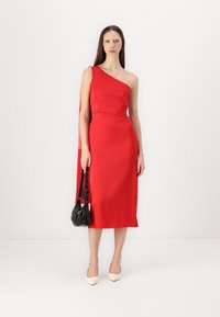 Robe asymétrique rouge, texture satinée, corsage ajusté, détail de cape fluide, longueur genou, associée à un sac à main noir et des chaussures crème.