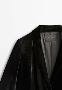 Blazer en velours noir avec un col cranté, texture douce, et une étiquette à l'intérieur. L'intérieur est doublé d'un tissu lisse.