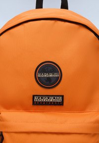 Zaino in tessuto arancione con un patch rotondo del logo e un'etichetta rettangolare del marchio, entrambi ben visibili sulla parte anteriore.