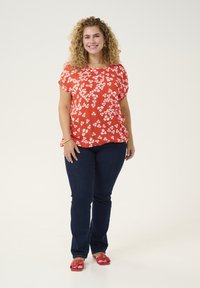 Rød blomsterbluse med korte ærmer, parret med mørkeblå jeans. Røde sandaler med remme og dekorative hardware detaljer.