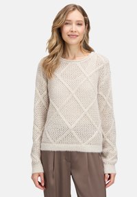 Hellbeige, locker gestrickter Pullover mit Rautenmuster, gerippten Bündchen und Saum sowie einer weichen, flauschigen Textur. Über braunen Hosen getragen.