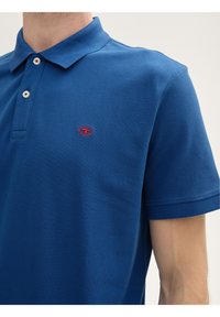Niebieska koszulka polo z teksturowanej tkaniny, z klasycznym kołnierzykiem, trzema guzikami i małym czerwonym logo po lewej stronie piersi.