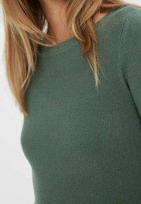 Femme avec de longs cheveux blonds portant un haut à manches longues ajusté en côtes vert foncé, vu des épaules jusqu'au milieu du torse.