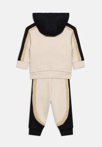 Παιδικό σετ φόρμας δύο τεμαχίων με κουκούλα και παντελόνι που ταιριάζουν, μπεζ με μαύρες και καφέ καμπύλες ρίγες στο πλάι.