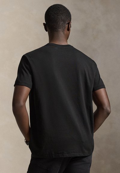 Άνδρας που φαίνεται από πίσω φορώντας ένα απλό μαύρο κοντομάνικο μπλουζάκι και μαύρο παντελόνι, στέκεται μπροστά σε μπεζ φόντο.