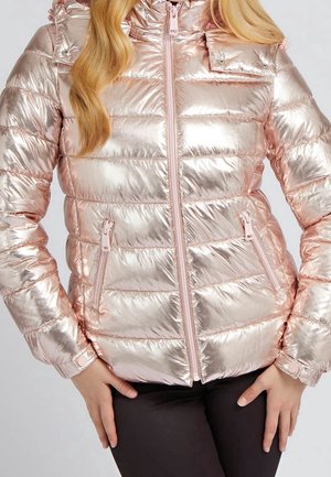 Błyszcząca, różowozłota pikowana kurtka zapinana na suwak z kieszeniami na zamek, noszona przez osobę z długimi blond włosami i czarnymi spodniami.