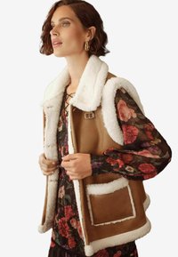 Gilet in finta shearling marrone con colletto e rifiniture bianche, dotato di tasche laterali e una chiusura in metallo, indossato sopra un vestito floreale.