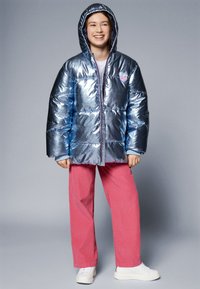 Doudoune bleue brillante avec capuche, ornée de rectangles cousus, d'un patch en forme de cœur et d'une fermeture éclair. Assortie à un pantalon rose et des chaussures blanches.