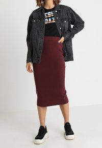 Schwarze Jeansjacke über einem schwarzen T-Shirt mit buntem Text, kombiniert mit einem bordeauxfarbenen Bleistiftrock und schwarzen Sneakers.