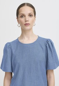 Bluză de culoare albastră deschisă, cu mâneci scurte și voluminoase, decolteu rotund și linii texturate verticale. Material moale cu accente subtile de strălucire.