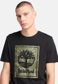 Camiseta de algodón negra con un gran logo de Timberland en verde y negro dentro de un diseño rectangular con un patrón de fondo similar al camuflaje.