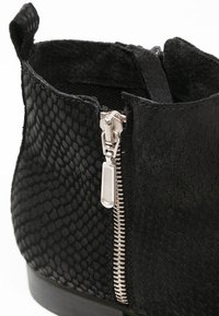 Primer plano de una bota negra de tobillo con una superficie texturizada y una cremallera plateada en el lateral, que muestra un diseño elegante y una excelente artesanía.