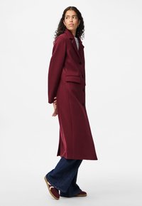 Μπορντό μακρύ παλτό με γιακά με εγκοπή, μαύρα κουμπιά και πλαϊνές τσέπες, συνδυασμένο με καμπάνα τζιν παντελόνια και κόκκινα αθλητικά παπούτσια. Ομαλή υφή.