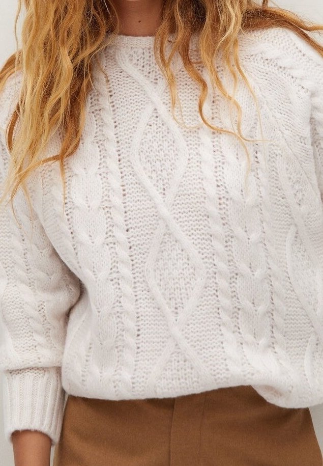 Femme portant un pull en maille torsadée blanc avec des motifs texturés et des manches longues, associé à un pantalon marron taille haute.