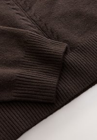 Dark brown sweater detail showing soft, textured fabric, ribbed cuffs, and a smooth surface with a subtle pattern along the seam.