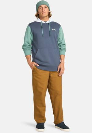 Hoodie mit türkisfarbenen Ärmeln und einem marineblauen Körper, ausgestattet mit einer Kängurutasche, getragen mit beigen Hosen und marineblauen Schuhen. Eine gestrickte Mütze rundet den Look ab.