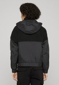 Svart jakke med en myk, teksturert øvre del og en glatt nedre del; har hette og elastiske mansjetter for en tettsittende passform.