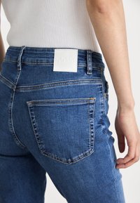Personne portant un jean en denim bleu avec une étiquette blanche sur la taille et un haut blanc côtelé, vue de dos de la taille à la cuisse.