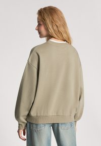 Jasnooliwkowy sweatshirt z okrągłym dekoltem, ściągaczami na rękawach i u dołu, noszony na bladii niebieskich dżinsach. Miękka tkanina o luźnym kroju.