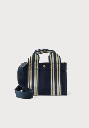Lille navy blue tote bag med brede, vertikale cremefarvede og navy stribede håndtag samt en justerbar navy skulderrem, sat mod en hvid baggrund.