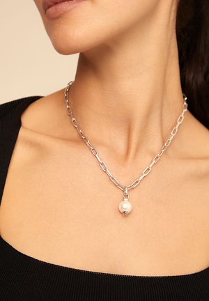 Collar de cadena de plata con un único colgante de perla blanca, llevado en el cuello de una mujer sobre una blusa negra de escote cuadrado.