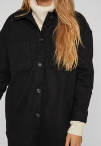 Črna zimska jakna s kragnjo, ki ima prsno žepek, debele gumbe in sproščen kroj. Manšete odkrijejo pleteno pulover pod njo.