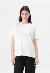 White cotton T-shirt with short sleeves, featuring a graphic design of dots forming a bra shape on the front. Smooth texture and relaxed fit.