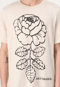 Beige t-skjorte med korte ermer som har et stort svart rosegrafikk med detaljerte kronblader og blader, samt "marimekko"-logoen.