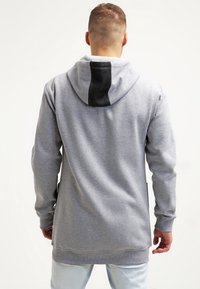 Šedá mikina s kapucí a kontrastním černým panely na kapuci, vyrobená z měkké tkaniny, má uvolněný střih a boční kapsy.