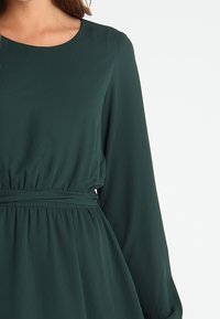 Dunkelgrünes Kleid mit rundem Ausschnitt, langen Ärmeln und geraffter Taille. Hergestellt aus einem glatten, leichten Stoff. Schlichte Designelemente.