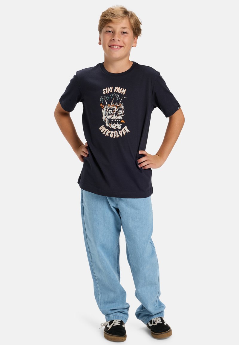 Granatowa bawełniana koszulka z grafiką czaszki i kolorowymi akcentami, zestawiona z jasnoniebieskimi luźnymi dżinsami i czarnymi trampkami.