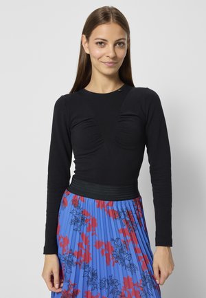 Zwarte top met lange mouwen en een geplooide V-vorm, gecombineerd met een blauwe geplooide rok met een rood bloemenpatroon en een zwarte tailleband.