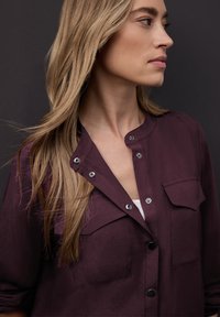 Chemise bordeaux boutonnée avec un col arrondi, deux poches poitrine et des boutons argentés en accent, fabriquée en tissu lisse.