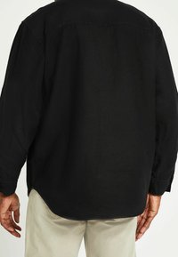 Czarna koszula z długim rękawem o prostym kroju. Posiada kołnierzyk zapinany na guziki, teksturowaną tkaninę i zaokrąglony dół. Model nosi jasne spodnie.