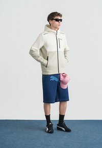 Νέος άνδρας που φοράει μπεζ φούτερ με φερμουάρ Puma, τζιν σορτς με λογότυπο Puma, μαύρα αθλητικά παπούτσια, μαύρες κάλτσες, γυαλιά ηλίου, κρατώντας ροζ καπέλο στο πλάι.