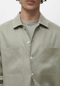 Svetlo zelena lanena srajca z izrezanim ovratnikom, dvema sprednjima žepoma in belimi gumbi. Gladka tekstura, sproščen kroj, primerna za plastenje.