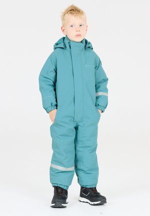 Combinaison de neige imperméable turquoise avec fermeture éclaire sur le devant, capuche, poignets élastiques et bandes réfléchissantes. Associée à des bottes d'hiver noires, se tenant sur un fond blanc.