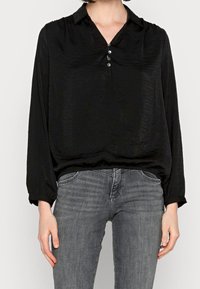 Maglietta nera a manica lunga in tessuto leggero con colletto e tre bottoni frontali, abbinata a jeans in denim grigi.