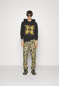 Felpa nera con stampa grafica gialla e testo, abbinata a pantaloni a fantasia in nero, giallo e bianco. Completata da scarpe nere e oro.