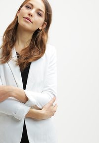 Hvit blazer med skreddersydd design, lett stoff og jakkeslag. Modellen står med armene krysset mot en ensfarget bakgrunn.