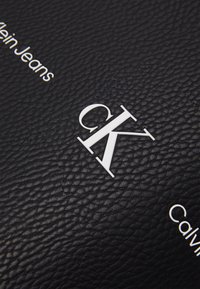 Czarna, teksturowana skóra z białym wytłoczonym logo "CK" oraz napisem "Calvin Klein Jeans". Powierzchnia ma wyraźny wzór ziarna.