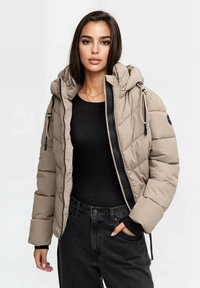 Beige Daunenjacke mit Kapuze, ausgestattet mit einem Frontreißverschluss, elastischen Bündchen und einem gesteppten Design. Getragen über einem schwarzen Oberteil mit dunkler Jeans.