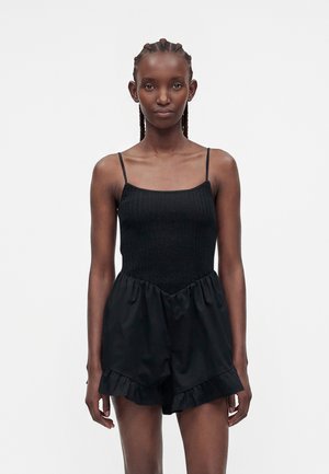 Jeune femme aux cheveux tressés portant une combinaison noire sans manches avec fines bretelles et short à volants, debout sur un fond blanc uni.