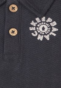 Marineblaues Polohemd mit einem strukturierten Fischgrätmuster, drei Holzknöpfen und einem weißen gestickten Logo-Design auf der Vorderseite.