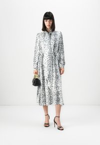Eine Frau steht in einem weißen Midikleid mit schwarzen Punkten, hält eine kleine schwarze Handtasche und trägt schwarze High-Heels vor einem schlichten weißen Hintergrund.